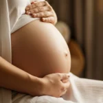 عکس واقعی از شکم بزرگ زن باردار در اواخر سه ماهه سوم، با دست هایی که به آرامی شکم را نوازش می کنند، در نور طلایی و گرم.